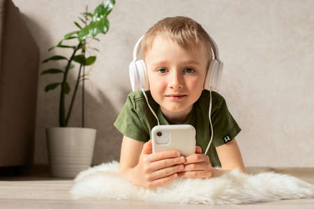 A child boy with a phone is lying on the floor with headphones watching cartoons, listening to music, playing games, talking on the phone.の写真素材