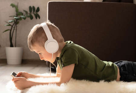 A child boy with a phone is lying on the floor with headphones watching cartoons, listening to...