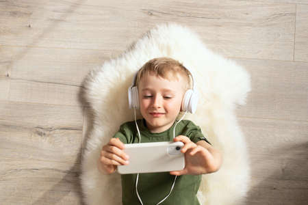 A child boy with a phone is lying on the floor with headphones watching cartoons, listening to music, playing games, talking on the phone. Top viewの写真素材