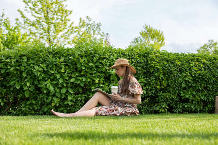 A young brunette girl is sitting on the grass in the park, drinking coffee and relaxing, working on a tablet, shopping online, learning.の写真素材