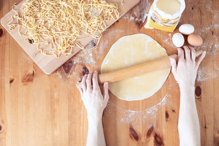 The process of cooking homemade noodles, spaghetti, pasta. On a wooden table, there is dough, a rolling pin, eggs, a board and a knife. Top view. The girl prepares homemade noodles, rolls out the dough and slices it thinly.の写真素材