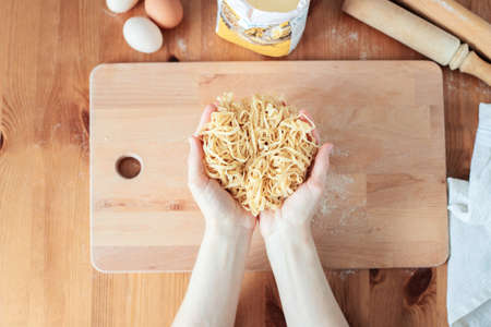 The process of cooking homemade noodles, spaghetti, pasta. On a wooden table, there is dough, a rolling pin, eggs, a board and a knife. Top view. The girl prepares homemade noodles, rolls out the dough and slices it thinly.の写真素材
