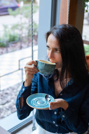 A brunette girl in a blue shirt and white trousers sits in a cafe by the window and drinks tea