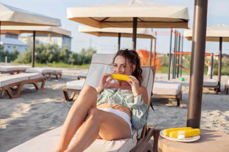 A young brunette girl is lying on a sunbed on the beach and eating boiled corn. Summer food. Summer...