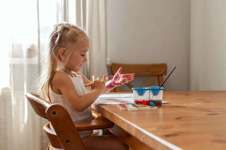 A little blonde girl is drawing at the table with paints. The girl plays, indulges, laughs, paints herself with paint. Children's emotionsの写真素材