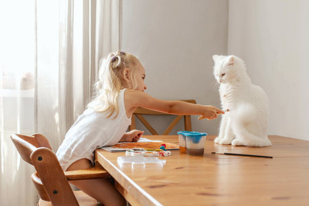 A little blonde girl draws at the table with paints with a white cat. The girl plays with the cat, indulges, laughs, paints the cat and herself with paint. Children's emotionsの写真素材