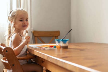 A little blonde girl is drawing at the table with paints. The girl plays, indulges, laughs, paints herself with paint. Children's emotionsの写真素材