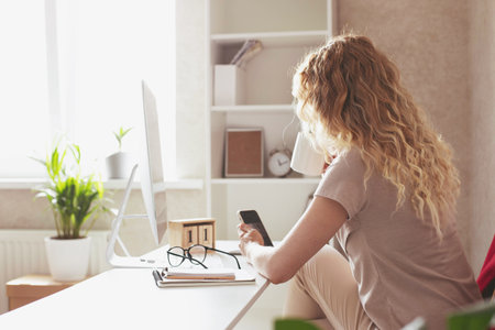 A young blonde girl works in a modern office. Drinks tea, talks on the phone, looks at the phone.の写真素材