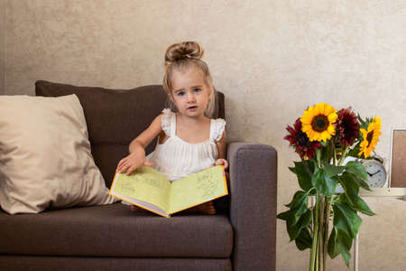 A little girl is sitting in a chair and looking at a book. Children's leisure. Sunflowers in the interior.の写真素材