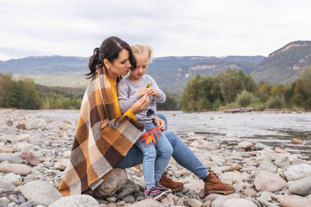 Mom and daughter are sitting on the bank of a mountain river in the forest in autumn, wrapped in a blanket, talking, having fun, throwing pebbles into the water.の写真素材