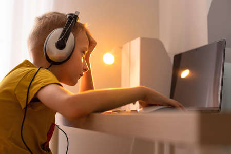 The boy is sitting at the table with headphones on and playing computer games. There is a laptop and...