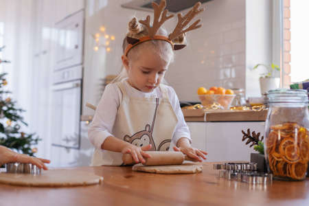 A little girl in an apron and reindeer horns with her mother rolls out the dough on the table for Christmas cookies. The interior of the Christmas kitchen. Family holiday Cooking.の写真素材