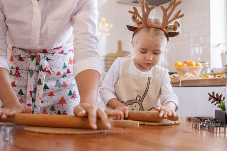 A little girl in an apron and reindeer horns with her mother rolls out the dough on the table for Christmas cookies. The interior of the Christmas kitchen. Family holiday Cooking.の写真素材