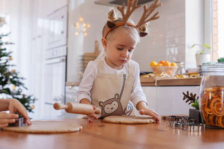 A little girl in an apron and reindeer horns with her mother rolls out the dough on the table for...