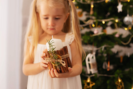 A little girl is holding a Christmas candle decorated with cinnamon sticks, a sprig of rosemary and a dried orange on the background of a Christmas tree. New Year's interior.の写真素材