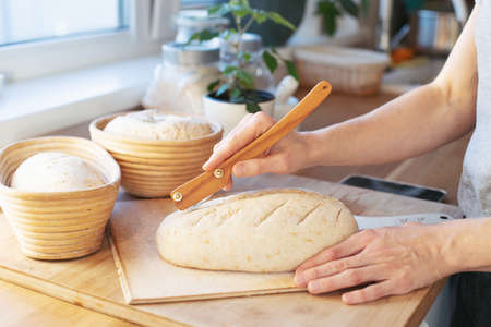 Preparations of sourdough Bread in rasstoechnye rattan baskets are on the table. The girl makes incisions with a knife and puts it in the oven to bake. The interior of a home kitchen. Homemade sourdough bread with your own hands.の写真素材