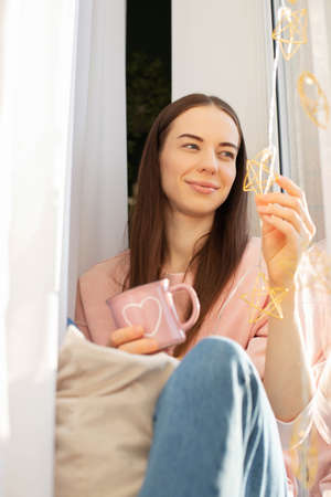 A young girl is sitting on the window holding a mug of tea, coffee, hot drinks, looking out the window, dreaming, resting.の写真素材