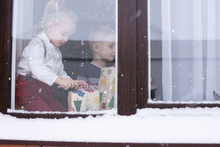 Children sit on the window in winter on Christmas holidays with gifts in their hands, a Christmas tree is decorated against the background.の写真素材
