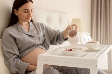 A pregnant brunette girl in pajamas is having breakfast in b ed with granola with yogurt, drinking...