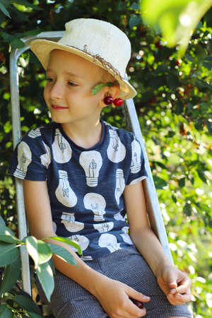 A boy in the village in the garden in a straw hat is sitting on a stepladder eating cherries. Spend summer holidays with his grandmother in the village.の写真素材