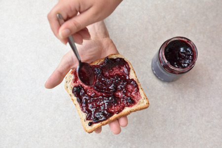 Blueberry jam with a spoon put on a piece of bread, which lies in the hand.