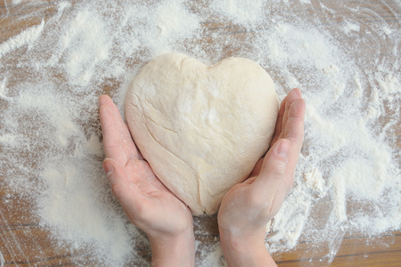 Raw dough in the shape of a heart and a touch of hands to it. Hands gently touch the raw dough in the form of heart. Close-up.の写真素材