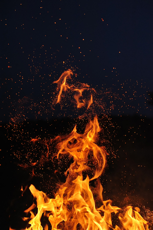 Flame of a burning campfire with sparks against a dark blue sky.