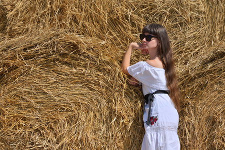 A brunette girl in a white dress and glasses, against a background of a large haystack from round bales.の写真素材