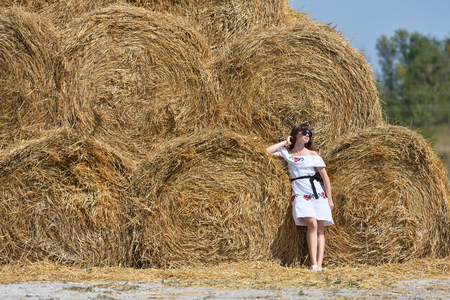 A brunette girl in a white dress and glasses, against a background of a large haystack from round bales.の写真素材