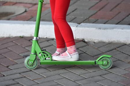A little girl in red pants and white sandals rides a scooter.