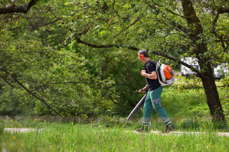 A male worker blows leaves and debris from the sidewalk with a blower.