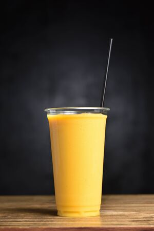 Summer cocktail of mango, banana, ice and juice in a plastic glass with a straw tube on a black background.の写真素材
