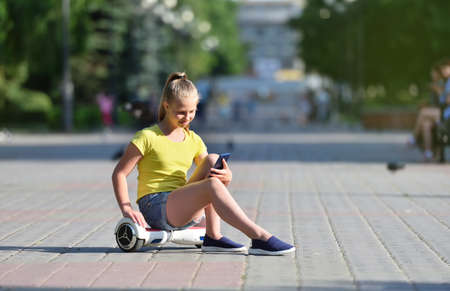 Happy girl child looking at her smartphone and sitting on a hoverboard in the parkの写真素材