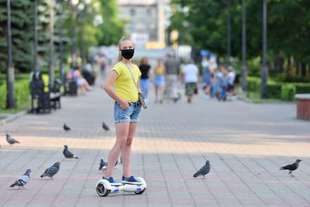 Girl child riding a hoverboard in a protective mask on the background of the park during the COVID-19 pandemicの写真素材