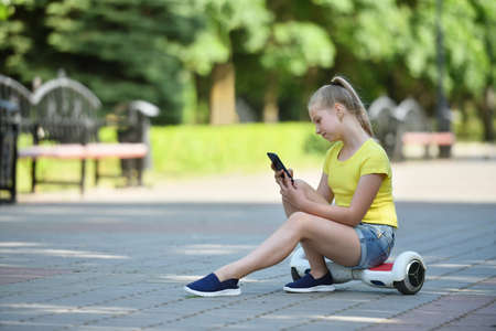 Girl child looks carefully at a smartphone and sits on a hoverboard in a parkの写真素材