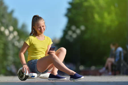 Smiling girl child reads message in a smartphone while walking in the park on a hoverboardの写真素材