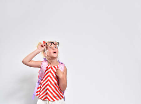 Schoolgirl 7 years old looks up in surprise raising her glasses on a gray background, studio...