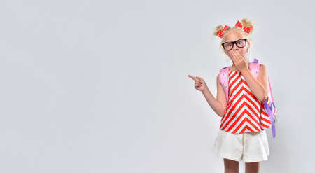 Schoolgirl 7 years old in glasses looks at the camera in surprise and shows her fingers to the sideの写真素材
