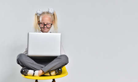Cute little girl 7 years old in glasses using a laptop on the background of a gray wall with a copy...