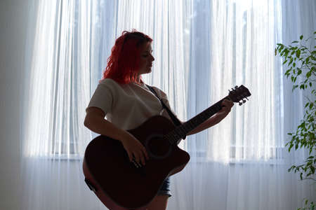 A teenager girl silhouetted against the background of the window plays the guitarの写真素材