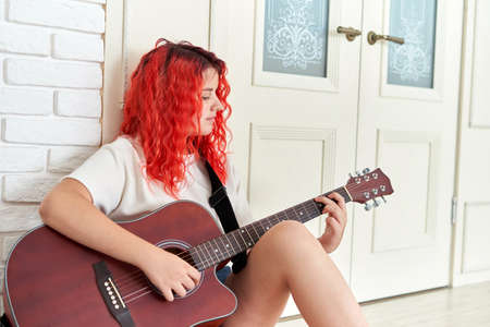 Cute teenage girl with red hair sits on the floor and plays a red guitarの写真素材