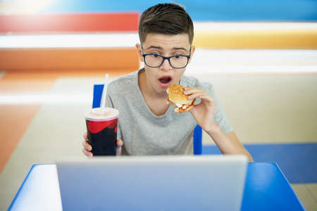 Caucasian white teenager eating burger and looking surprised at laptop