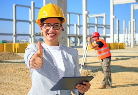 Happy beautiful female architect with tablet on construction site. She is smiling and satisfied with her job, giving thumb up looking at camera, behind her construction worker with measuring deviceの写真素材