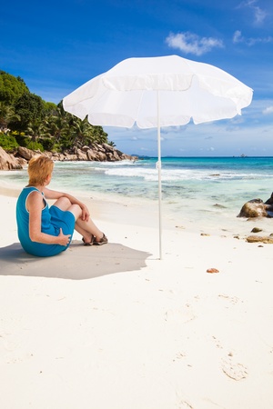 woman sitting under a white sunshade in the sand of a tropical beach with green palms and brown granite rocks, a turquoise sea and a blue sky with white clouds, Seychelles, La Digueの写真素材