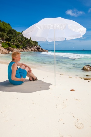 woman sitting under a white sunshade in the sand of a tropical beach with green palms and brown granite rocks, a turquoise sea and a blue sky with white clouds, Seychelles, La Digueの写真素材