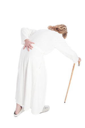 invalid old woman in a white bath robe with pain in the back walking with a stick and pressing one hand against her aching backの写真素材