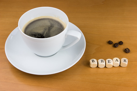 a white cup of coffee on a brown wooden table with some coffee beans and five letter cubes showing the word break. Concept for have a coffee break.の写真素材