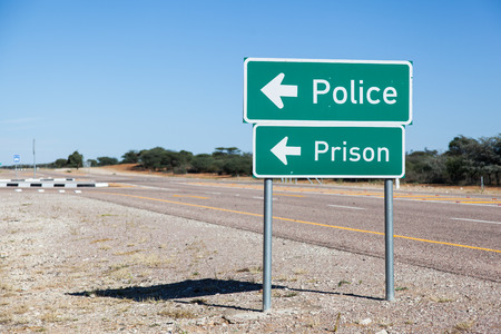 two green metal at a tar road signs showing the way to police and prison concept for the result of crimeの写真素材