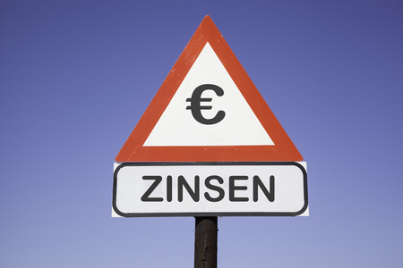 White road warning triangle with black  exclamation point and red frame on  a wooden mast in front of a blue sky. A second rectangular sign warns in german about interest rateの写真素材