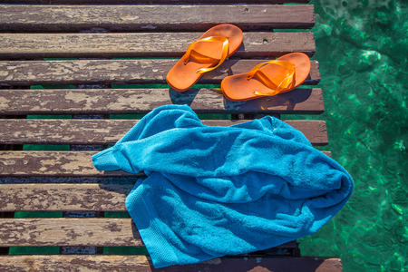 pair of orange flip-flops and a blue towel on the wooden planks of a jetty over the seaの写真素材